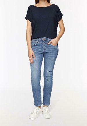 Femme portant un haut bleu marine foncé à manches courtes, un jean skinny bleu clair déchiré et des baskets blanches, debout avec une main dans la poche.