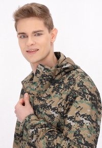 Jakna s kamuflažnim uzorkom u nijansama zelene, smeđe i crne. Ima visok ovratnik, zatvaranje na zip, te prilagodljive manšete. Vodootporni materijal.