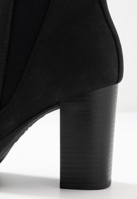 Schwarze Wildleder-Stiefelette mit Blockabsatz und strukturiertem Oberflächenmaterial sowie elastischen Seiteneinsätzen für einfaches Anziehen.