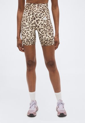 Personne portant un short de cycliste taille haute à imprimé léopard, des chaussettes blanches montantes et des baskets rose clair, debout devant un fond uni.