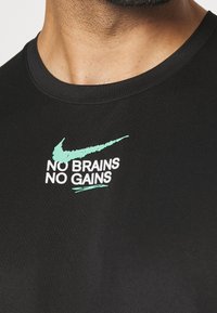 Černé bavlněné tričko s krátkými rukávy a žebrovaným výstřihem. Na přední straně se nachází velký Nike swoosh v mátově zelené barvě a text "ŽÁDNÉ MOZKY, ŽÁDNÉ ZISKY" v bílé barvě.