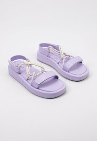 Sandalias de plataforma de color púrpura claro con suelas planas, una tira frontal ancha y cordones entrelazados que presentan acentos contrastantes en amarillo y blanco.