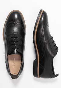 Chaussures oxford en cuir noir avec fermeture à lacets, détails brogués et un petit talon en caoutchouc. Elles présentent une surface lisse et des accents de semelle extérieure beige.