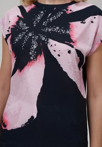 Kortsleeve, marineblå og rosa topp med floral design. Materialet er mykt med en glatt tekstur, dekorert med metalliske nagler på blomsten.