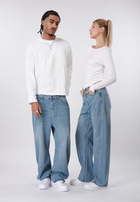 Δύο μοντέλα φορούν καθημερινά σύνολα: ένα λευκό τοπ με μακριά μανίκια και φαρδιά τζιν παντελόνια ανοιχτού μπλε χρώματος. Σημαντικές λεπτομέρειες περιλαμβάνουν άνετη εφαρμογή και λευκά αθλητικά παπούτσια.