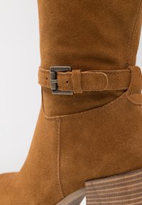 Bota de ante marrón hasta la rodilla con una correa de hebilla lateral, acabado texturizado y un tacón de madera apilada. Detalle de costura suave visible.
