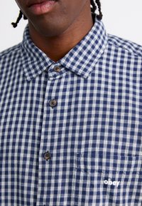 Camisa de cuadros en azul marino y blanca con cuello clásico, que presenta botones marrones y un logo "obey" en blanco en el bolsillo.