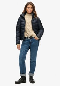 Marineblauwe gewatteerde jas met een aansluitend ontwerp, crèmekleurige kabelgebreide trui, blauwe recht-toe-rechtaan jeans en zwarte veterschoenen.