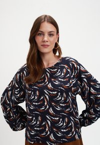 Kobieta z długimi brązowymi włosami, nosząca złote kolczyki w kształcie kół oraz granatową bluzkę w białe i miedziowe abstrakcyjne wzory.