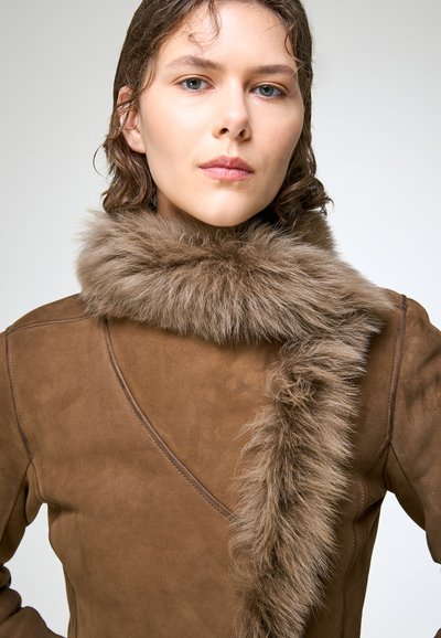 Jeune personne aux cheveux courts et bruns portant un manteau en daim marron avec un large col en fourrure, regardant droit devant avec une expression neutre.