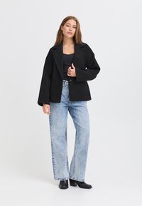 Giovane donna in piedi, che indossa un blazer nero, una maglietta nera, jeans a gamba larga azzurro chiaro e stivali neri con tacco alla caviglia, su uno sfondo bianco semplice.