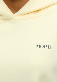 Άτομο που φοράει ένα ανοιχτό κίτρινο φούτερ με την επιγραφή "MOP D" τυπωμένη με μικρά μαύρα γράμματα στο στήθος.