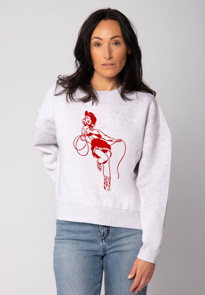 Sweatshirt gris à coupe décontractée, avec un graphique rouge représentant une femme portant un chapeau et tenant un lasso, associé à un jean en denim.