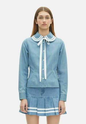 Jonge vrouw in een lichtblauwe denim blouse met knopen en een witte strik, en een geplooide denim rok met witte strepen.
