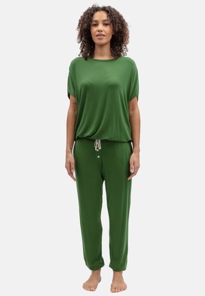 Femme debout pieds nus portant un haut vert ample à manches courtes et un pantalon assorti à cordon, sur fond blanc.