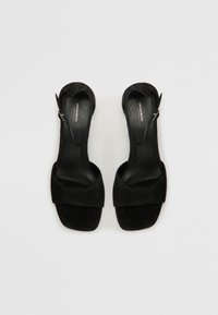 Fekete velúr slingback szandál, széles elülső pánttal és karcsú sarokkal. Simább belső bélés és állítható bokapánt a tökéletes illeszkedés érdekében.