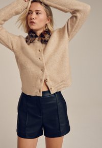 Cardigan court beige texturé avec un col en fausse fourrure et de gros boutons, associé à un short en cuir noir.
