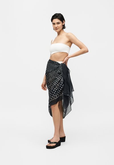 Γυναίκα που φοράει λευκό ασύμμετρο crop top, μαύρη φούστα με σχέδια και μαύρα σανδάλια πλατφόρμα, στέκεται με το χέρι στη μέση σε απλό φόντο.