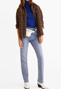 Chemise à carreaux brune par-dessus un col roulé bleu, associée à un jean bleu clair et des bottines blanches, avec un sac banane blanc.