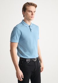 Tricou polo albastru deschis, cu mâneci scurte, având guler și închidere cu două butoane, asortat cu blugi negri și o curea neagră.