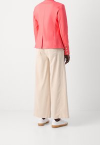 Veste ajustée corail avec une fente unique et des poignets boutonnés, assortie à un pantalon beige à jambes larges et des baskets blanches avec semelles en caoutchouc.