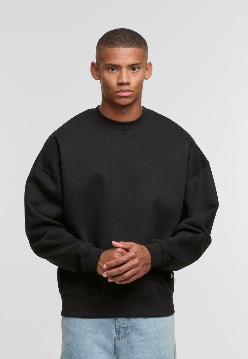 Schwarzer übergroßer Sweatshirt mit Rundhalsausschnitt, gerippten Bündchen und Saum, aus weichem Material. Kombiniert mit hellblauen Jeans.