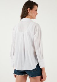 White button-up shirt with long sleeves, lightweight fabric, and a slightly curved hem, paired with denim shorts.