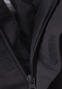 Juodos spalvos užtrauktuku užsegamas striukės vidus su lygiu audiniu, Velcro detalėmis ir kontrastuojančiais užtrauktuko elementais saugiam uždarymui.