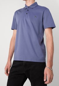 Polo tričko ve tlumené purpurové barvě, s límečkem, propínáním na tři knoflíky a nášivkou s logem na hrudi. Krátké rukávy a hladká textura látky.