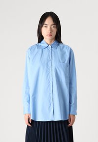 Light blue button-up shirt with a pointed collar, long sleeves, and a chest pocket, made from smooth fabric with a relaxed fit.