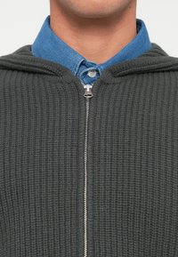 Felpa grigia con zip e superficie testurizzata, dotata di cappuccio. Indossata sopra una camicia button-up azzurra chiara con colletto. Dettagli in zip argentati.
