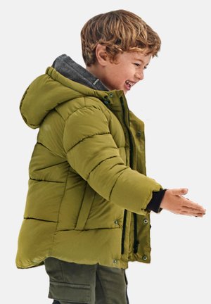 Giacca puffer verde con cappuccio, dotata di sezioni trapuntate, una cerniera frontale e chiusure a pressione. Indossata sopra un maglione grigio e pantaloni scuri.