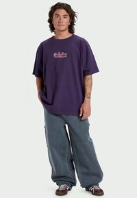 Paarse oversized t-shirt met een ronde hals, voorzien van een klein geborduurd logo op de borst, gecombineerd met losvallende blauwe spijkerbroeken en bruine schoenen.