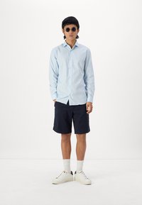 Lichtblauw overhemd met lange mouwen, gecombineerd met donkerblauwe shorts en witte sneakers. Accessoires bestaan uit ronde zonnebrillen.