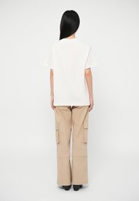 Γυναίκα με μακριά μαύρα μαλλ�ιά φοράει λευκό κοντομάνικο πουκάμισο και μπεζ παντελόνια cargo, στέκεται μπροστά από ένα απλό λευκό φόντο.