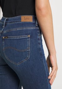Mörkblå denimjeans med en slät yta, som har en framträdande brun läderlapp med "Lee"-logotypen i midjan och stiliga sömmar på fickorna.