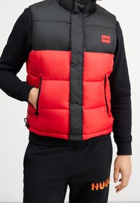 Personne portant un gilet matelassé noir et rouge avec l'étiquette « HUGO », un sweat-shirt noir et un pantalon noir avec un texte « HUGO » en flammes orange sur la jambe gauche.