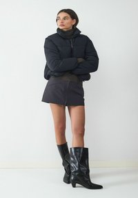 Czarna kurtka puchowa z wysokim kołnierzem, dopasowany krój, w połączeniu z ciemną spódniczką mini w paski i eleganckimi wysokimi, czarnymi botkami.