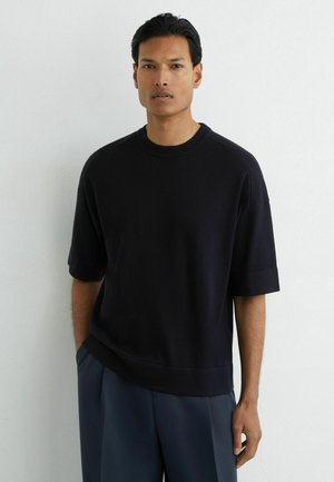 Mann mit kurzen schwarzen Haaren, der einen schwarzen Pullover mit kurzen Ärmeln und marineblaue Hose trägt, steht an einer weißen Wand.