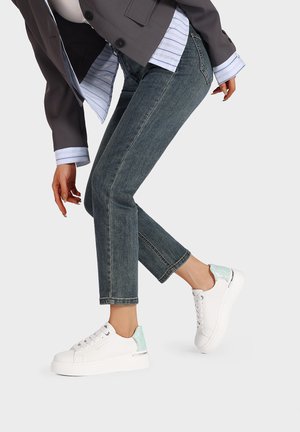 Sneaker bianche con suola spessa, caratterizzate da un accento verde menta sul tallone e occhielli argentati. Indossate con jeans blu a vita alta.