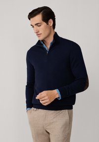 Mann in einem navyblauen Sweatshirt mit Viertelreißverschluss über einem blauen Hemd und beigen Hosen, schaut nach unten, mit einer Hand in der Tasche und der anderen in der Nähe der Taille.