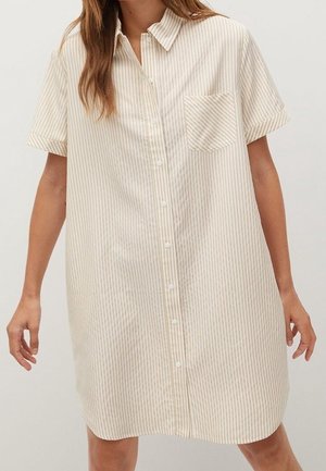 Femme portant une robe-chemise ample, longueur genoux, manches courtes, avec des rayures verticales beige et blanches et une poche poitrine.