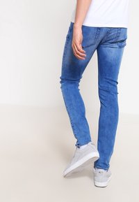 Jeans de denim azul de corte ajustado con un lavado desgastado, que presentan sutiles bolsillos traseros y se combinan con zapatillas grises sobre un fondo claro.