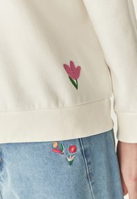 Sudadera de algodón blanca con un tulipán bordado en rosa en el costado. Jeans de mezclilla azul con bordados florales coloridos cerca de la cinturilla.