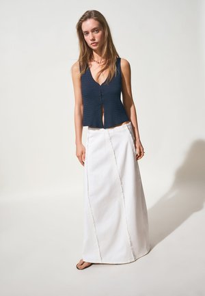 Giovane donna che indossa un top smanicato blu navy con bottoni e una gonna lunga bianca, in piedi scalza davanti a uno sfondo chiaro e uniforme.