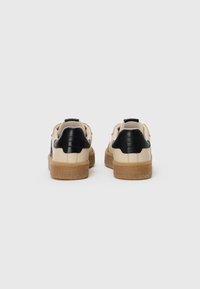 Baskets avec une tige en cuir beige, des accents de talon texturé noirs et une semelle en caoutchouc marron, présentant un design minimaliste sans logos visibles.