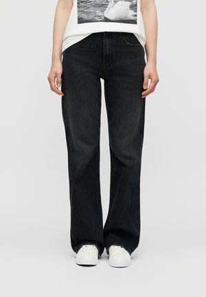 Osoba oblečená v voľných čiernych džínsoch s rozstrapkanými lemami a bielych teniskách, v kombinácii s bielou košeľou s potlačou labuťovej fotografie.