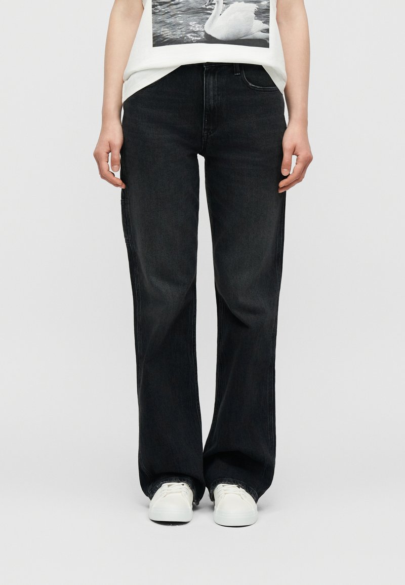 Άτομο που φοράει φαρδιά μαύρα τζιν με ξεφτισμένα τελειώματα και λευκά αθλητικά παπούτσια, συνδυασμένα με λευκό πουκάμισο με φωτογραφικό τύπωμα κύκνου.