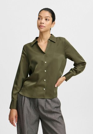 Blusa di raso verde oliva con maniche lunghe, chiusura frontale con bottoni e colletto. Texture liscia con un leggero lucido, abbinata a pantaloni fantasia.