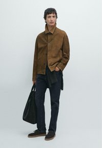 Giovane uomo in piedi contro uno sfondo semplice, indossa una giacca in suede marrone, jeans scuri, scarpe marroni e tiene una borsa tote nera.